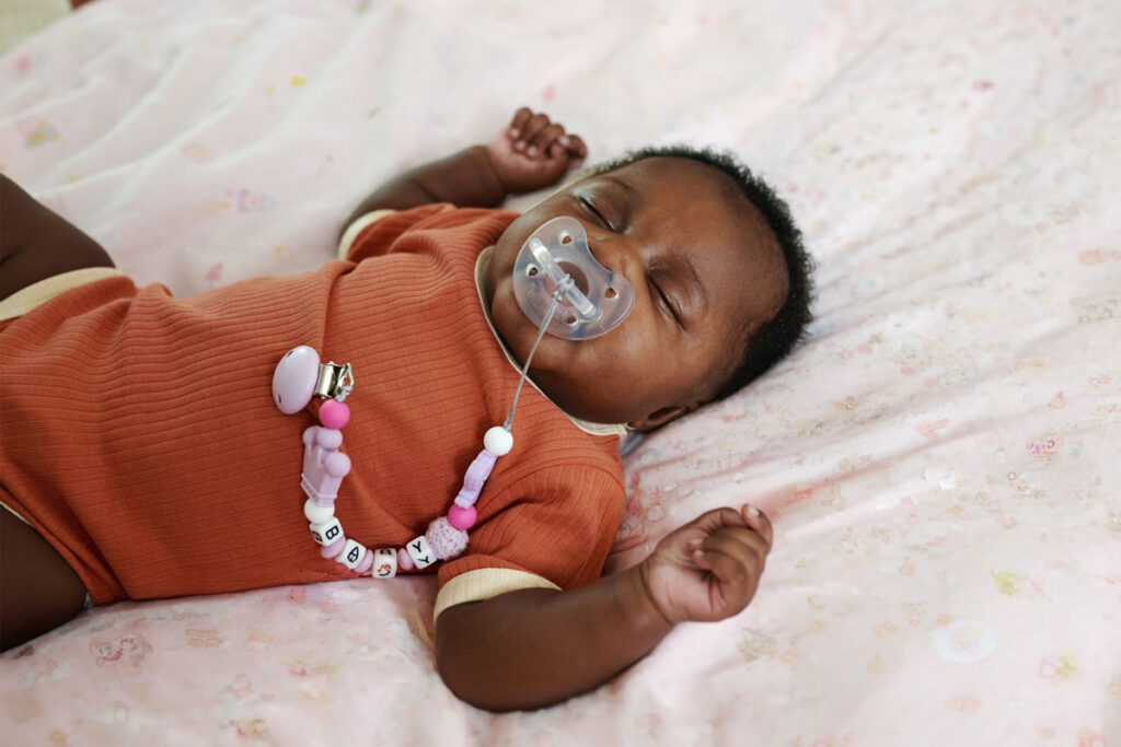 Sleeping baby with pacifier clip lying on pink blanket.