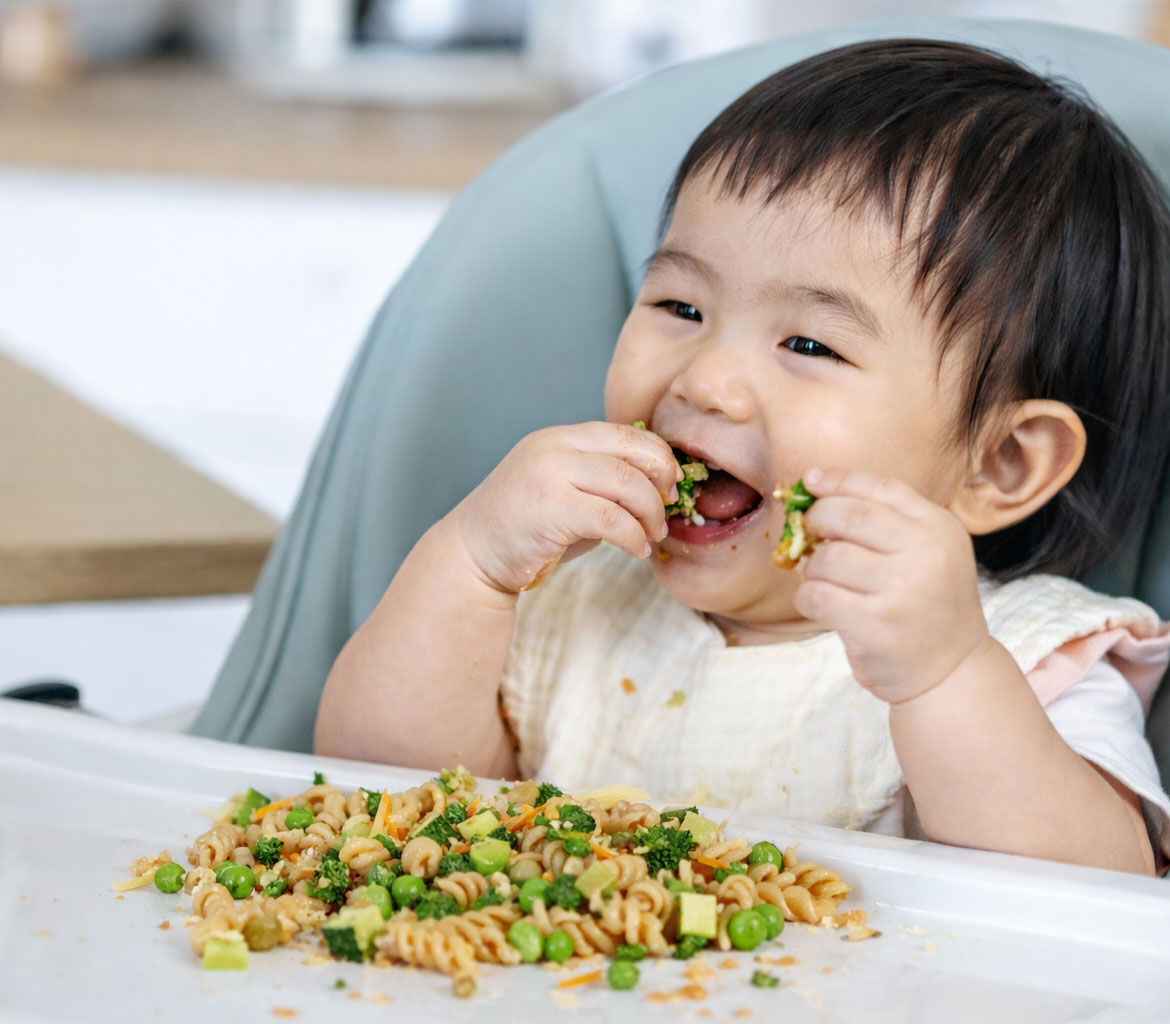Baby happily eating pasta and vegetables in high chair
