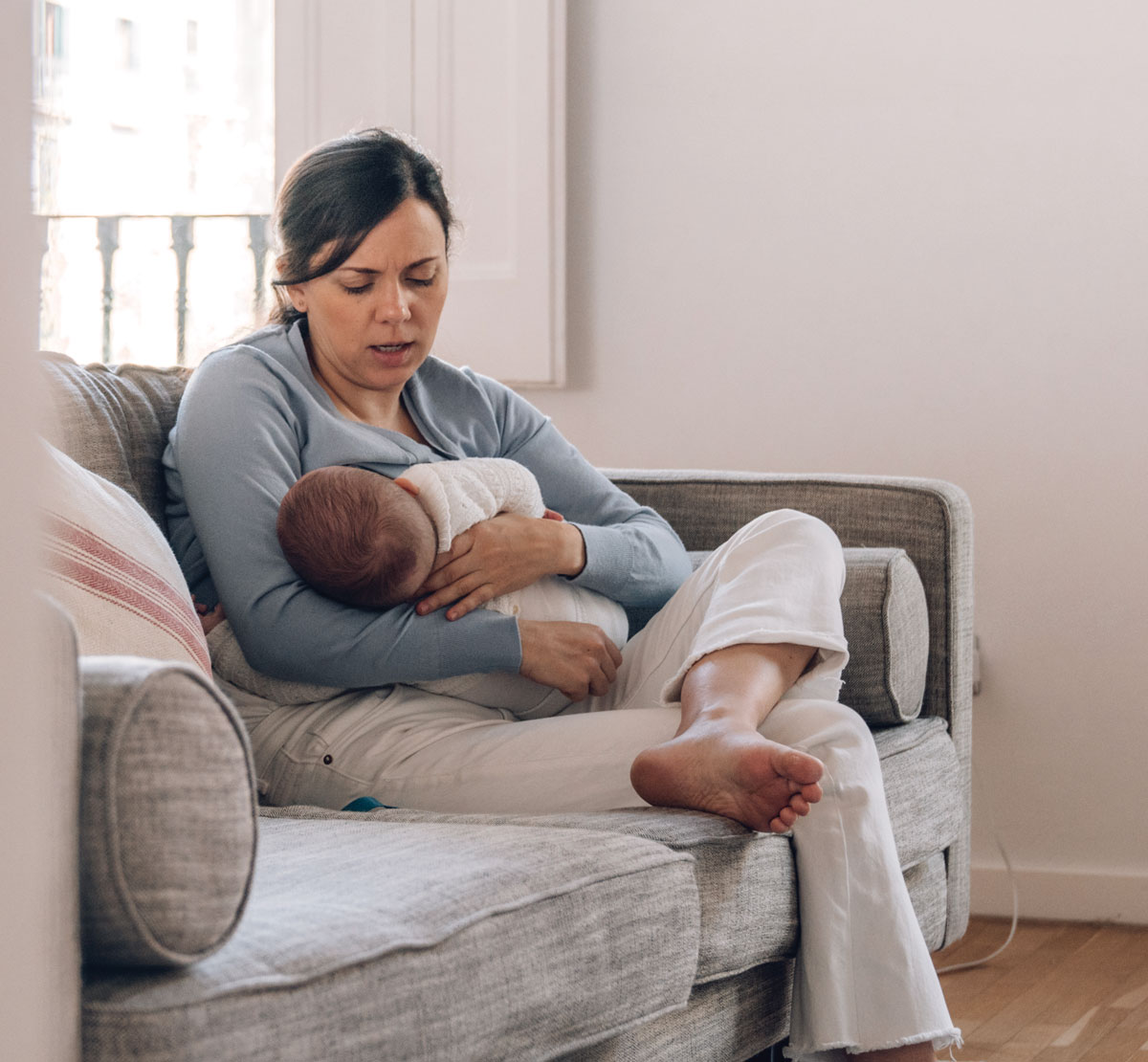 Mother experiencing breastfeeding discomfort while nursing her baby on a couch.