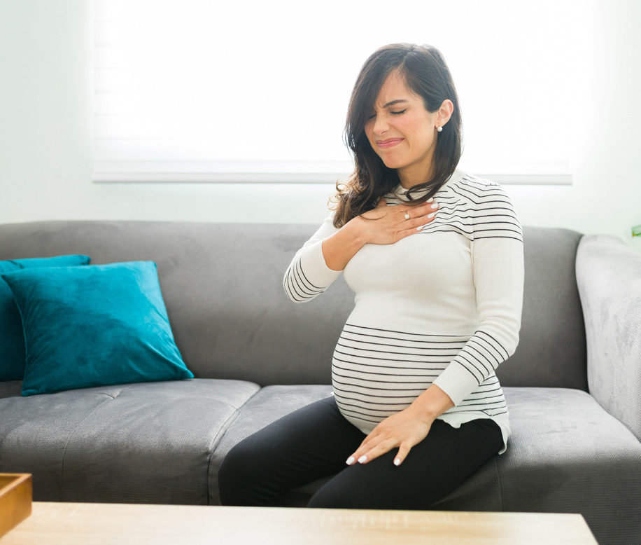 Pregnant woman experiencing heartburn, sitting on grey couch.